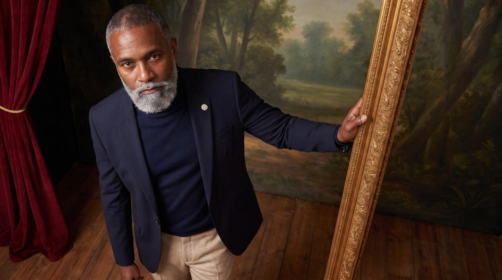 Man wearing a navy blazer and turtleneck beside an ornate gold frame and red velvet curtains